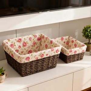 May include: Two woven brown storage baskets with floral fabric lining. The fabric features pink roses on a cream background. The baskets are rectangular and sit on a white surface.