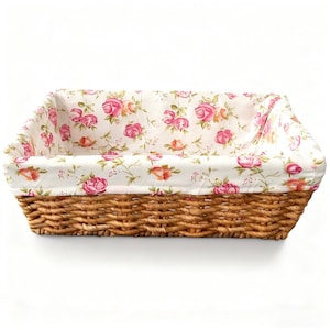 May include: Rectangular woven basket with a floral fabric lining. The basket is made of natural brown woven material. The fabric lining features pink and orange roses on a white background. The basket is empty.