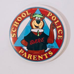 May include: A circular white badge with a cartoon bear wearing a green hat and a black shirt with the word "DARE." The badge has a red and blue border with the words "SCHOOL POLICE PARENTS."