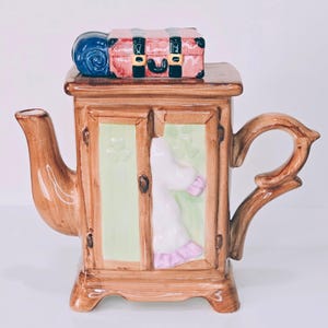 May include: A whimsical ceramic teapot shaped like a wardrobe, with a trunk and rolled blanket on top. The wardrobe doors reveal a white dress with pink trim. The teapot is brown with green and pink accents.
