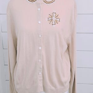 May include: A light beige cardigan with a decorative collar and button-up front. The collar is embellished with white and gold beading. A cross-shaped design with gold and white beads adorns the front of the sweater. The long-sleeved cardigan is displayed on a mannequin.