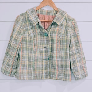 May include: A vintage plaid jacket in pastel colors of green, yellow, and pink. The jacket has a collared neckline, three buttons, and three-quarter sleeves. The jacket is hanging on a wooden hanger against a white background.