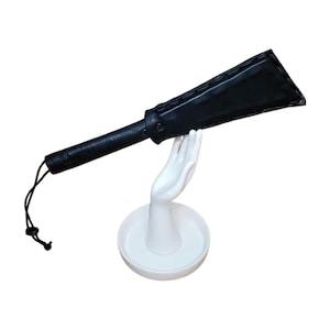May include: A black leather paddle with a textured handle and a loop at the end. The paddle is displayed on a white hand-shaped stand with a circular base. The paddle's head is a trapezoid shape.