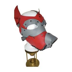 May include: A red and gray leather pup mask with a pointed ear design. The mask features a snout and is adorned with silver rivets. The mask is displayed on a white mannequin head.