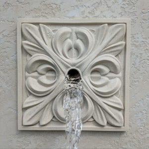 May include: A white concrete wall fountain with a decorative, ornate design. Water flows from a spout in the center of the design.