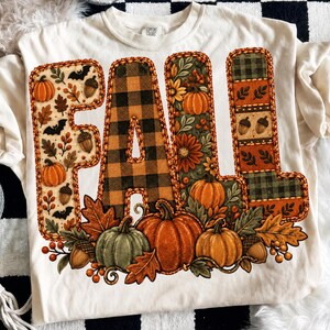 May include: A cream-coloured t-shirt featuring the word FALL in large block letters. The letters are filled with patterns of pumpkins, acorns, bats, leaves, and plaid. Below the letters are illustrations of three pumpkins and acorns.