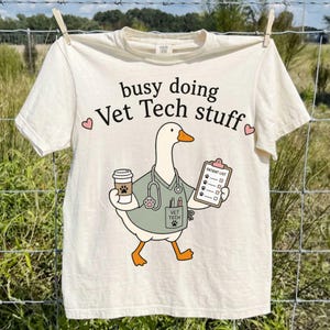 May include: A cream-colored t-shirt featuring a graphic of a white goose dressed in a sage green veterinary scrub top with a stethoscope, holding a coffee cup and a clipboard labeled Patient List. The text busy doing Vet Tech stuff is printed above.