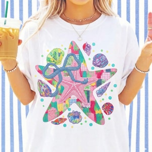 May include: White t-shirt featuring a colorful star design with a pink starfish, seashells, and a blue bow. The star is made of patchwork fabric in various colors. The design includes small blue dots.
