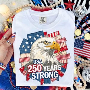 Pode incluir: Camiseta branca com um design patriótico de águia e o texto "USA 250 YEARS STRONG". O gráfico incorpora elementos da bandeira americana, incluindo estrelas e listras. A camisa é da marca Comfort Colors, tamanho M-M-M.