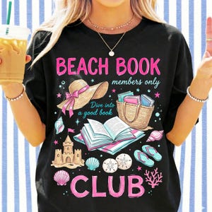 May include: Black t-shirt featuring a beach-themed design with a straw hat, books, and seashells. The text on the shirt reads "Beach Book Club" in pink, along with "Dive into a good book". The design evokes a seaside theme.