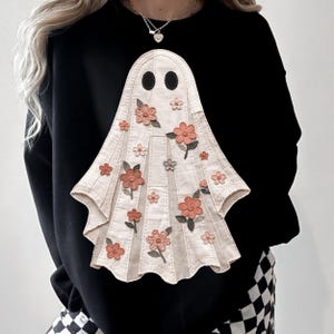 May include: A black long-sleeved sweatshirt featuring a large, central appliqué of a cream-coloured ghost. The ghost is decorated with various embroidered peach and pink flowers with green leaves, and has two solid black oval eyes.