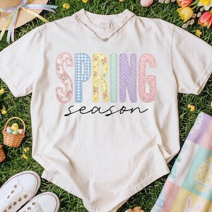 May include: Cream-colored t-shirt with the words "SPRING season" in colorful, patterned letters. The word "SPRING" is in a mix of floral, gingham, and polka dot patterns. The shirt is on a bed of green grass.