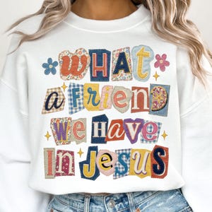 以下が含まれることがあります： 「WHAT A FRIEND WE HAVE IN JESUS」の文字がカラフルなパッチワーク風にデザインされた白いスウェットシャツ。花柄やギンガムチェック柄、星や花のアクセントが特徴。カジュアルで着心地の良いスタイルです。