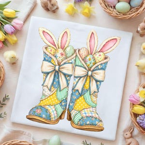 Puede incluir: Camiseta blanca con una colorida ilustración de dos botas de patchwork llenas de huevos de Pascua y orejas de conejo. Las botas tienen grandes lazos color crema. El diseño incorpora patrones florales azules, amarillos y verdes, perfecto para la Pascua.