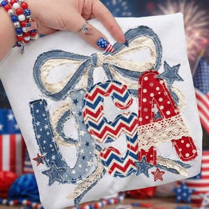 May include: White square fabric with the letters "USA" appliqued in red, white, and blue patriotic patterns. The letters are embellished with lace and stars. A large denim bow and stars complete the design.
