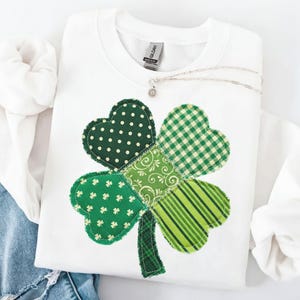 Può includere: Felpa bianca girocollo con un grande trifoglio applicato. Il trifoglio è composto da quattro foglie a forma di cuore in vari motivi verdi, tra cui pois, quadretti e righe. Una collana d'argento con un piccolo pendente è appoggiata sulla maglia.