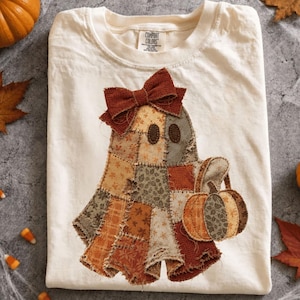 Puede incluir: Una camiseta de color crema con un gráfico central de un fantasma hecho de varios parches de tela marrón, naranja y bronceado. El fantasma tiene ojos ovalados oscuros, un gran lazo marrón en la cabeza y sostiene una pequeña calabaza a juego.
