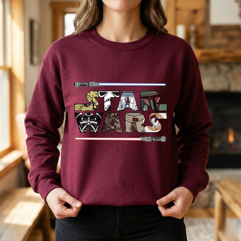 Puede incluir: Sudadera burdeos con la inscripci&oacute;n "STAR WARS" en una fuente colorida y estilizada. Las letras est&aacute;n dise&ntilde;adas con personajes y elementos ic&oacute;nicos de Star Wars. Dos sables de luz, uno azul y otro rojo, flanquean el texto.