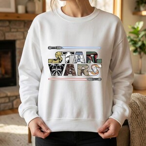 Puede incluir: Sudadera blanca con la inscripci&oacute;n "STAR WARS" en un dise&ntilde;o de letras de bloque colorido. Las letras est&aacute;n rellenas de varios gr&aacute;ficos de personajes y s&iacute;mbolos. Dos sables de luz, uno azul y otro rojo, est&aacute;n colocados encima y debajo del texto.