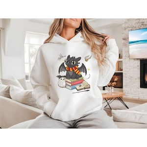 May include: White hooded sweatshirt featuring a cartoon dragon with a red and yellow scarf, sitting on a stack of books. An owl, a wand, and other magical elements are also depicted. The sweatshirt is a comfortable, casual style.