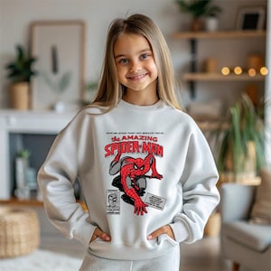 Puede incluir: Sudadera gris claro con un gr&aacute;fico de c&oacute;mic de Spider-Man. El gr&aacute;fico incluye las palabras "The Amazing Spider-Man" en rojo y blanco, con un dibujo de Spider-Man. La sudadera tiene mangas largas.