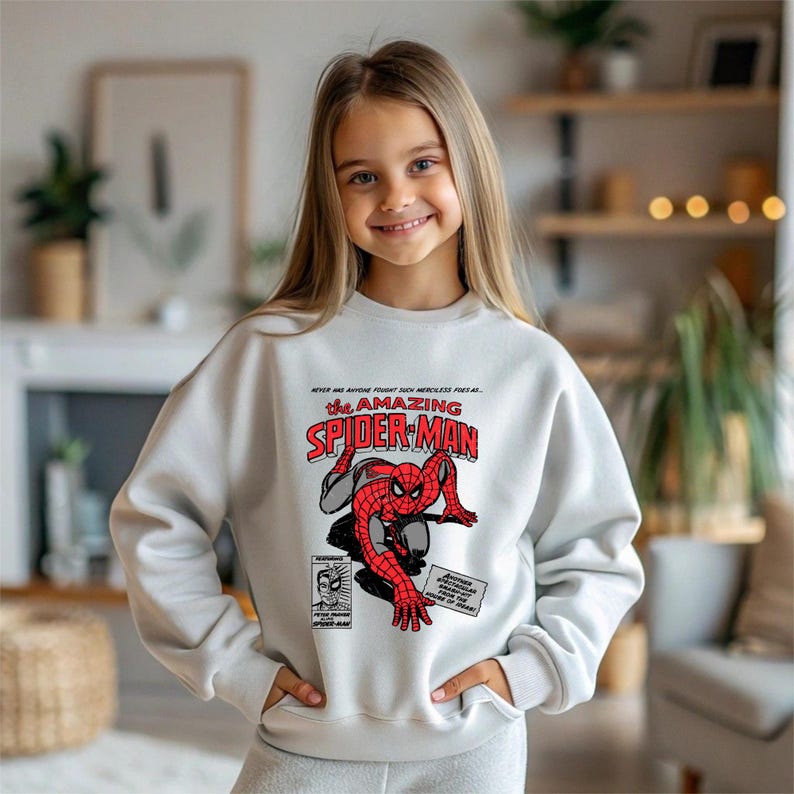Puede incluir: Sudadera gris claro con un gr&aacute;fico de c&oacute;mic de Spider-Man. El gr&aacute;fico incluye las palabras "The Amazing Spider-Man" en rojo y blanco, con un dibujo de Spider-Man. La sudadera tiene mangas largas.