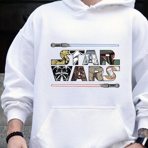 Puede incluir: Sudadera con capucha blanca con el texto "STAR WARS" en un dise&ntilde;o de letras en bloque colorido. El dise&ntilde;o incluye im&aacute;genes de personajes y sables de luz. La sudadera tiene un bolsillo delantero y una capucha con cord&oacute;n.