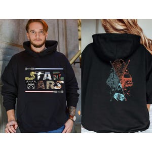Puede incluir: Sudadera con capucha negra con el texto "STAR WARS" en un dise&ntilde;o colorido y estilizado. La parte delantera de la sudadera muestra el texto con sables de luz. La parte trasera de la sudadera tiene un gr&aacute;fico de personajes de Star Wars.