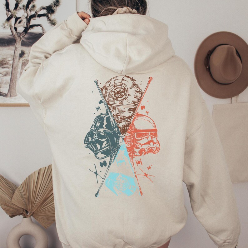 Puede incluir: Sudadera con capucha color crema con un gr&aacute;fico de Star Wars en la espalda. El dise&ntilde;o incluye ilustraciones del casco de Darth Vader, un casco de Stormtrooper y la Estrella de la Muerte, con detalles en azul, rojo y marr&oacute;n.