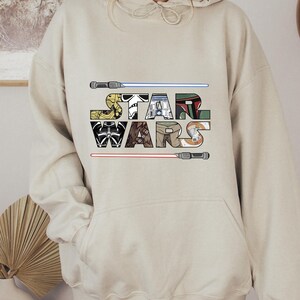 Puede incluir: Sudadera con capucha beige con la inscripci&oacute;n "STAR WARS", cada letra dise&ntilde;ada para parecerse a personajes y elementos de la franquicia Star Wars. Incluye sables de luz y dise&ntilde;os de personajes.