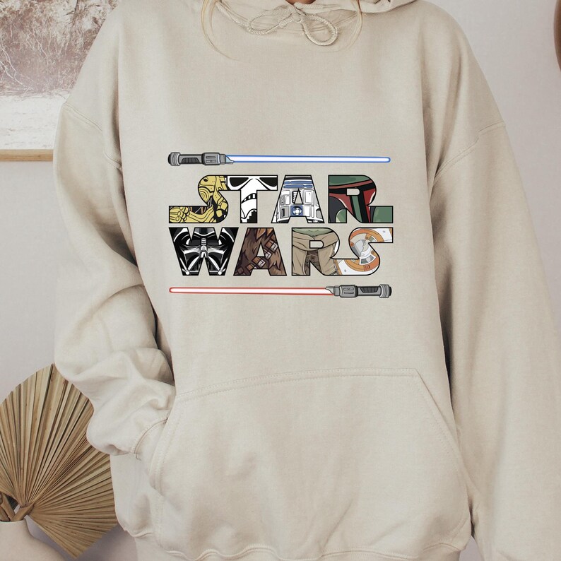Puede incluir: Sudadera con capucha beige con la inscripci&oacute;n "STAR WARS", cada letra dise&ntilde;ada para parecerse a personajes y elementos de la franquicia Star Wars. Incluye sables de luz y dise&ntilde;os de personajes.