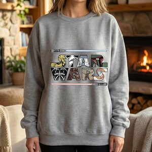 Puede incluir: Una sudadera gris con la inscripci&oacute;n "STAR WARS" en una fuente colorida y estilizada. Las letras est&aacute;n dise&ntilde;adas con im&aacute;genes de la franquicia Star Wars, incluyendo sables de luz y dise&ntilde;os de personajes.