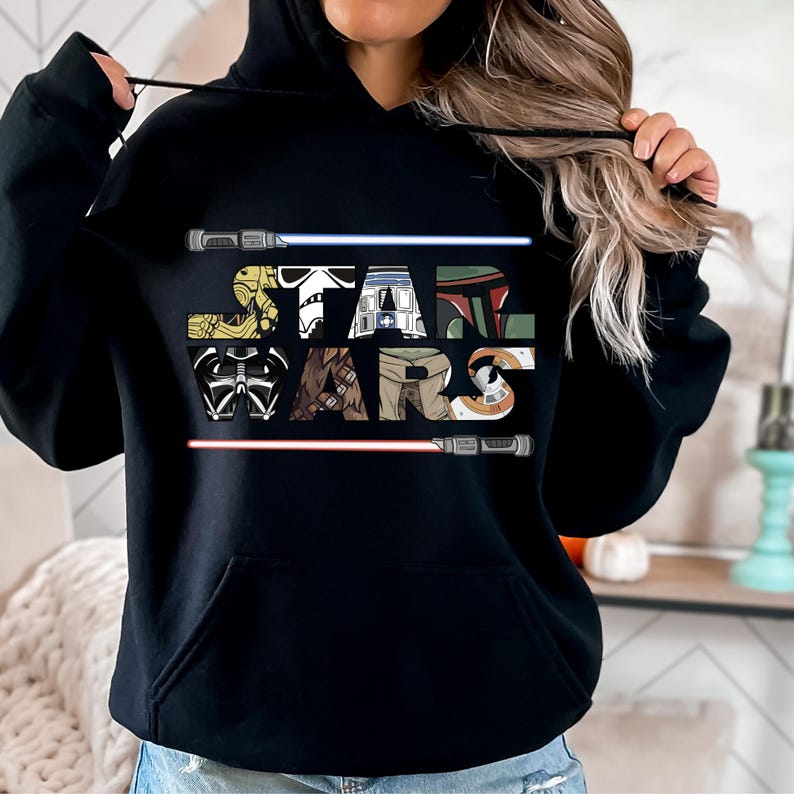 Puede incluir: Sudadera con capucha negra con la inscripci&oacute;n "STAR WARS", cada letra dise&ntilde;ada con personajes de Star Wars y sables de luz. El dise&ntilde;o incluye personajes como Darth Vader, Boba Fett y un Stormtrooper.