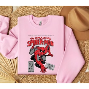 Puede incluir: Una sudadera rosa claro con un dise&ntilde;o de c&oacute;mic de Spider-Man. El gr&aacute;fico incluye el texto "the AMAZING SPIDER-MAN" en rojo y negro, con elementos de c&oacute;mic adicionales.