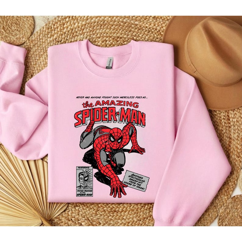 Puede incluir: Una sudadera rosa claro con un dise&ntilde;o de c&oacute;mic de Spider-Man. El gr&aacute;fico incluye el texto "the AMAZING SPIDER-MAN" en rojo y negro, con elementos de c&oacute;mic adicionales.
