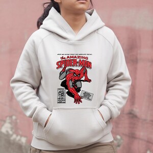 Puede incluir: Una sudadera con capucha blanca con un gr&aacute;fico de Spider-Man en rojo y negro. El dise&ntilde;o incluye las palabras "The Amazing Spider-Man" en rojo y blanco, junto con texto e ilustraciones de estilo c&oacute;mic.