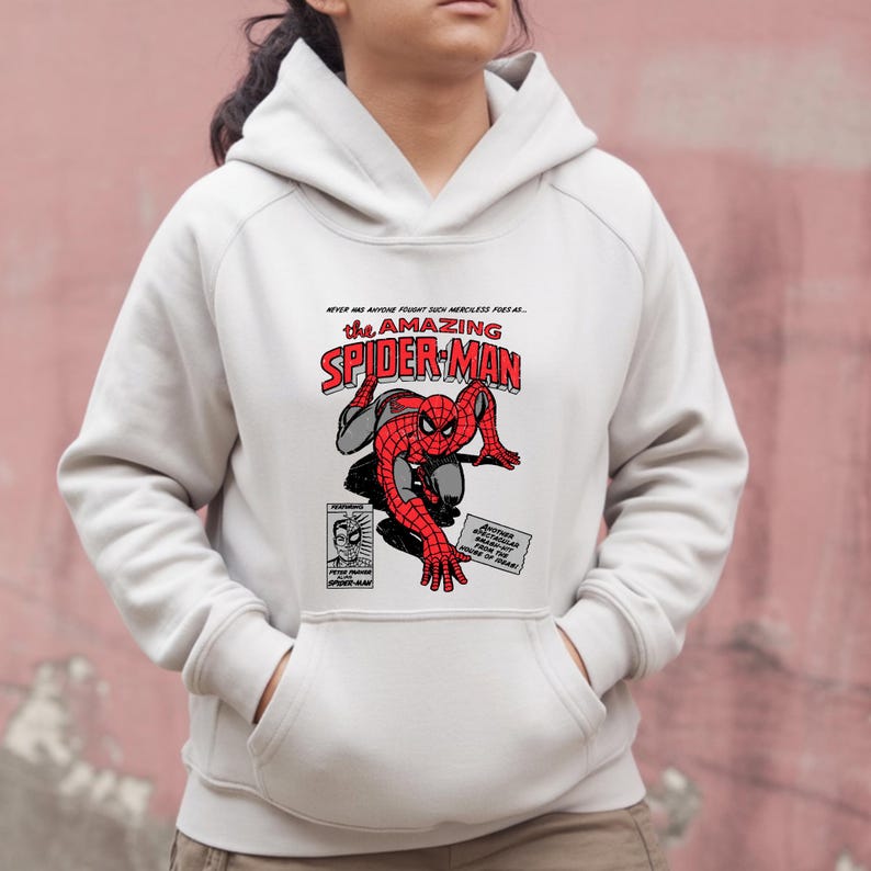 Puede incluir: Una sudadera con capucha blanca con un gr&aacute;fico de Spider-Man en rojo y negro. El dise&ntilde;o incluye las palabras "The Amazing Spider-Man" en rojo y blanco, junto con texto e ilustraciones de estilo c&oacute;mic.