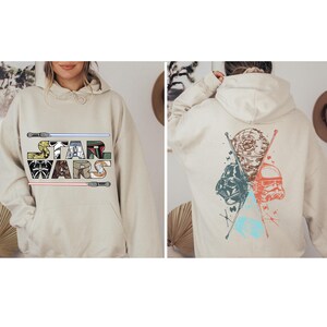 Puede incluir: Sudadera con capucha beige con el texto "STAR WARS" en un dise&ntilde;o colorido y en bloques en la parte delantera. La parte trasera muestra un gr&aacute;fico de Darth Vader, un Stormtrooper y la Estrella de la Muerte, con sables de luz y otros elementos.