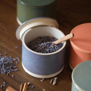 Op de afbeelding: Een set van vier cilindrische keramische potten met deksels, met matte afwerkingen in olijfgroen, terracotta, lichtblauw en maagdenpalmblauw. De open maagdenpalmblauwe pot bevat gedroogde lavendel en een houten lepeltje. Kaneelstokjes en gedroogde kruiden liggen verspreid.