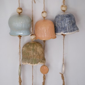 Puede incluir: Conjunto de carillones de cerámica en varios colores, incluyendo azul, melocotón, verde y azul marino. Cada carillón tiene forma de campana con líneas verticales y cuelga de una cuerda de hilo natural. Se ven cuentas decorativas y detalles de cerámica.