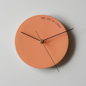 Puede incluir: Un reloj de pared redondo de color melocotón con manecillas negras y un centro de latón. El texto "THE JOY OF BEING" está impreso en negro en la parte superior. Un diseño minimalista para la decoración del hogar.