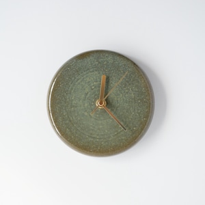 Puede incluir: Un reloj de pared redondo de cerámica con un esmalte verde apagado y manecillas doradas. El reloj tiene un diseño simple y minimalista sobre un fondo blanco. El diámetro del reloj es de aproximadamente 15 cm.