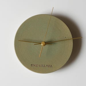 Puede incluir: Reloj de pared redondo verde salvia con manecillas y centro dorados. La esfera del reloj presenta la frase "TIME WILL TELL". Diseño minimalista, ideal para la decoración del hogar.