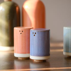 Puede incluir: Juego de saleros y pimenteros de cerámica en colores coral, azul y verde. Cada salero cilíndrico tiene una base blanca y tres agujeros. En el fondo, botellas de aceite a juego, sobre una superficie de madera.