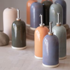 Puede incluir: Una colección de botellas de aceite de cerámica en varios colores, incluyendo naranja, verde y azul. Cada botella tiene un pico plateado y una base blanca. Las botellas están dispuestas sobre una superficie de madera clara.