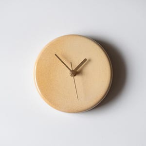 May include: A circular, beige wall clock with a minimalist design. The clock has a light beige face with gold-coloured hands and a second hand. The clock's face has a speckled texture. The clock is a simple, modern design.