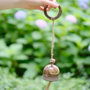 Pode incluir: Um sino de cerâmica castanho suspenso por um fio de juta e um anel de madeira. O sino tem uma forma arredondada com uma superfície texturizada. O fundo é um borrão de folhagem verde e roxa.