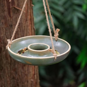 Puede incluir: Un comedero para pájaros de cerámica colgante con un diseño circular y una zona de alimentación central elevada. El comedero es de color verde apagado con un borde beige claro y está suspendido por tres cuerdas de cuerda natural. Se ve alpiste en el interior.