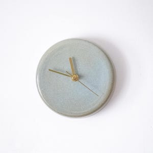 May include: A round, pale blue ceramic wall clock with gold-coloured hands and a central hub. The clock has a minimalist design and is mounted on a white wall. The clock's diameter is approximately 20 cm.