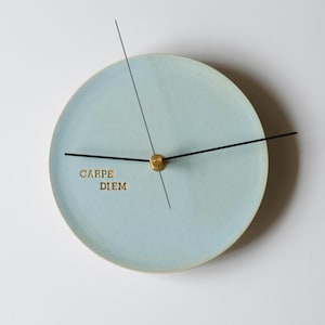 Puede incluir: Un reloj de pared redondo de color azul claro con manecillas negras y un centro dorado. La esfera del reloj tiene las palabras "CARPE DIEM" en letras doradas. El reloj tiene un diseño minimalista.